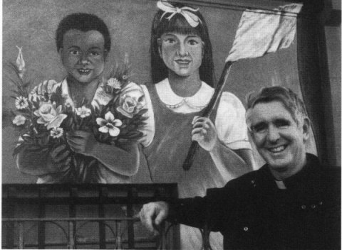 Picture of Father Isaacs in front of the Hispanic mural. the mural show two children the boy holding flowers and the girl a flag.